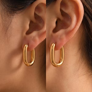 Könnte beinhalten: Ein Paar goldfarbene Creolen in länglicher ovaler Form mit einer glatten, polierten Oberfläche. Die Ohrringe sind als durchgehende Ringe mit einem Klappverschluss gestaltet und präsentieren eine schlichte, metallische Optik am Ohr.