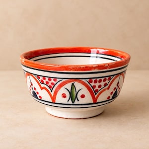 Op de afbeelding: Een keramische kom met een witte basis en een handgeschilderd patroon van oranje bogen, zwarte lijnen, rode stippen en groene bladvormige figuren. De rand is beschilderd in een effen oranje kleur en de kom heeft een ronde voet en opening.