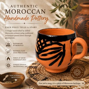Puede incluir: Una taza de cerámica color terracota con cuerpo redondeado y asa gruesa, decorada con patrones abstractos pintados en negro. La taza se apoya sobre una alfombrilla circular tejida. El texto en la imagen incluye AUTHENTIC MOROCCAN Handmade Pottery.