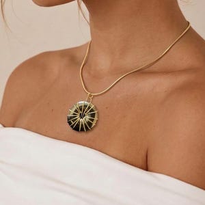 May include: A gold-toned snake chain necklace featuring a circular pendant. The pendant consists of a dark, round stone wrapped in thin gold-colored wire that radiates from the center to the outer edge, creating a distinct sunburst-like pattern.