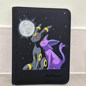 Può includere: Un raccoglitore nero con un'illustrazione dipinta di un Umbreon nero e un Espeon viola seduti sotto una luna piena e stelle. Il testo MintKeeper è stampato nell'angolo in basso a destra. Il raccoglitore ha una forma rettangolare.