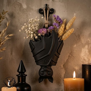May include: A matte black wall-mounted planter shaped like a hanging bat with a crescent moon and star engraving. The bat holds a bouquet of dried white baby's breath, purple statice flowers, and golden wheat stalks, suspended by a jute rope.
