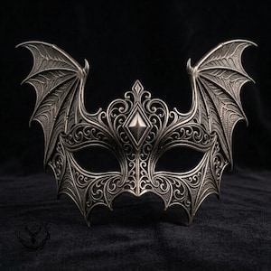 May include: A metallic silver masquerade mask featuring intricate filigree scrollwork and prominent bat-like wings extending from the sides. The center of the forehead displays a raised diamond-shaped accent, and the eye openings are almond-shaped.