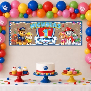 May include: A birthday party display featuring a rectangular banner with Paw Patrol characters and the text NICHOLAS 1ST BIRTHDAY, surrounded by a colorful balloon arch with paw print designs, a cake with a paw print, and cupcakes on stands.