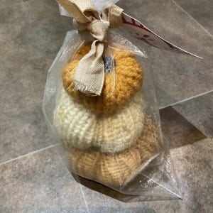 Op de afbeelding: Een doorzichtige cellofaan zak, vastgebonden met een jute lint en tule, bevat drie gestapelde, gebreide pompoenen in crème, geel en bruin. Een klein label is aan het lint bevestigd.