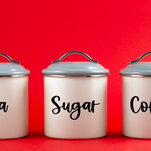 May include: Three white ceramic canisters with silver lids and black lettering. The canisters are labelled "Tea", "Sugar", and "Coffee".