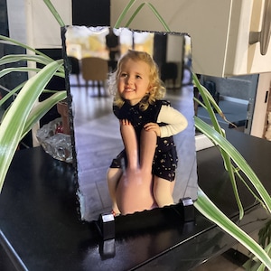May include: A black slate photo frame with a picture of a young girl sitting on a pink bunny. The frame has a black stand.