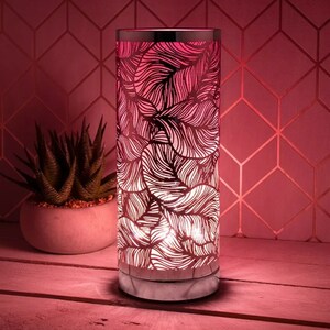 May include: A cylindrical lamp with a silver rim and base, featuring a leaf pattern cut into the pink-toned metal. The lamp is illuminated from within, casting a warm glow. A small succulent in a concrete pot sits nearby.