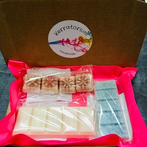 May include: A brown cardboard box containing handmade wax melts. The box is lined with pink tissue paper and includes various wax melt bars and cubes in shades of white, beige, and blue. The box has a circular sticker that says "Kerratorium Handmade".