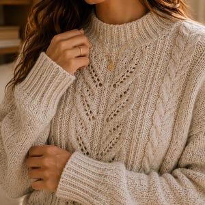 Puede incluir: Un suéter de punto trenzado en color beige con cuello alto, mangas largas con puños acanalados y un patrón texturizado de trenzas verticales y puntos calados. El tejido tiene una apariencia ligeramente moteada en todo el color neutro.