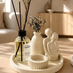 Puede incluir: Una bandeja redonda beige que contiene un difusor de varillas de vidrio con palitos negros y una etiqueta que dice AYU HOME FRAGRANCE FRESH AIR 120ml, un jarrón beige acanalado con flores secas, un portavelas beige acanalado y una figura de pareja.