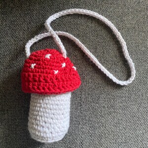 May include: A handmade crochet mushroom bag featuring a bright red cap with small white stitched accents and a white stem. The bag includes a long, thin white crocheted strap attached to the top of the mushroom cap for carrying purposes.