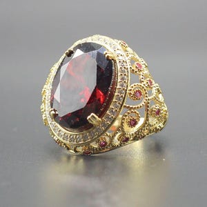 May include: A gold ring featuring a large oval-cut dark red garnet gemstone at the center, surrounded by a halo of small white stones. The band displays an intricate filigree pattern accented with tiny red stones along the sides of the setting.