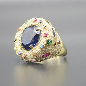 May include: A gold-toned ring featuring a large, oval-cut dark blue central stone surrounded by a halo of smaller, multi-colored gemstones including red, green, and clear stones. The band is wide and covered in tiny, pavé-set clear crystals.