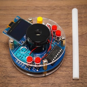 Puede incluir: Un conjunto de placa de circuito electrónico circular con una base azul, un zumbador negro, una pequeña pantalla rectangular y varios botones pulsadores rojos y amarillos. Los componentes están montados entre dos discos acrílicos transparentes.