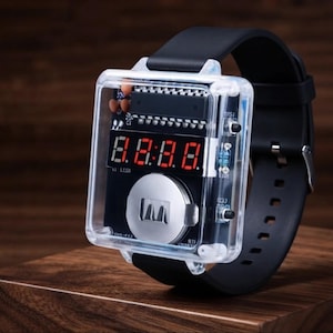 May include: A digital wristwatch featuring a clear, transparent square housing that reveals the internal black circuit board, electronic components, and a silver circular battery. The watch displays red digital numbers and has a black silicone strap.