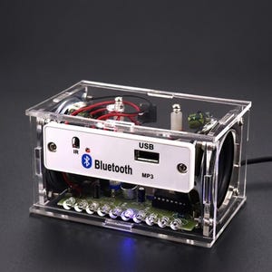 Puede incluir: Un kit de altavoz electrónico rectangular alojado en una caja de acrílico transparente. El panel frontal blanco presenta un puerto USB, un sensor infrarrojo y el texto Bluetooth y MP3. Una fila de pequeñas luces LED es visible en la base.