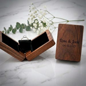 Puede incluir: Un joyero de madera marrón oscuro con tapa abatible, grabado con el texto Rose & Jack y la fecha 04.12.2027. La caja abierta revela un interior de terciopelo negro que sostiene un anillo plateado con una piedra preciosa redonda.