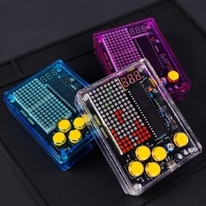 Puede incluir: Tres consolas de juegos electrónicas rectangulares con carcasas transparentes en azul, morado y transparente. Cada dispositivo cuenta con una pantalla LED de rejilla, un visor numérico de tres dígitos y cuatro botones circulares amarillos.