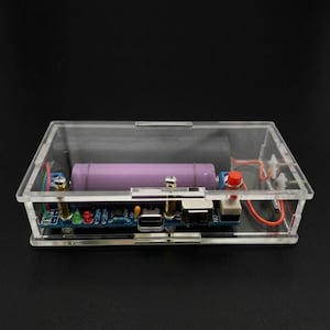 May include: A clear acrylic rectangular enclosure containing an electronic circuit board with a purple cylindrical battery, a red push button, a USB port, and various small components connected by red and green wires inside the transparent case.