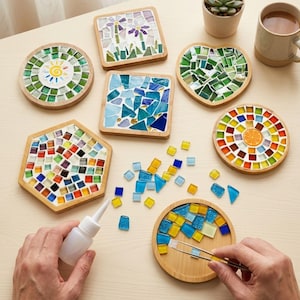 Puede incluir: Una colección de posavasos de madera con diseños de mosaico en varias formas, incluyendo circular, cuadrada, hexagonal y de corazón. Azulejos de vidrio sueltos, un frasco de pegamento y pinzas de metal están dispuestos para manualidades.