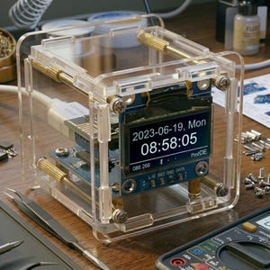 May include: A digital clock module housed in a clear acrylic cube frame with gold-toned metal standoffs. The screen displays the date 2023-06-19, Mon and the time 08:58:05. A white USB cable is connected to the blue circuit board inside the frame.