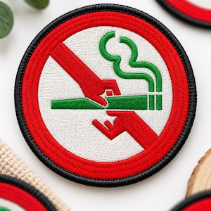 May include: A circular embroidered patch featuring a white background with a red border and a black stitched edge. The design displays a red prohibition symbol over a green cigarette being held by two red hands, with green smoke rising above.