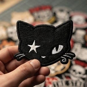 May include: A black embroidered cat head patch featuring a white star over one eye, a single white eye, and white stitched whiskers. The patch has a distinct black border and a flat, textured surface, showcasing detailed needlework throughout.