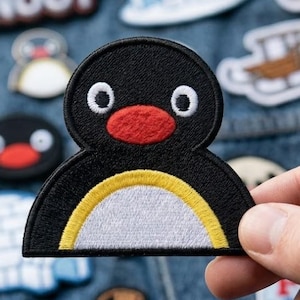 May include: An embroidered patch featuring a black penguin character with a red beak, white eyes, and a white belly outlined in yellow. The patch has a rounded top and a flat bottom edge, showcasing detailed stitching and a clean, defined border.