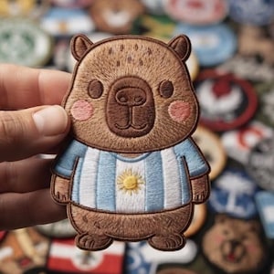 May include: An embroidered patch featuring a brown capybara with pink cheeks wearing a light blue and white striped shirt with a yellow sun emblem in the center, resembling the national flag of Argentina, with detailed brown thread stitching.