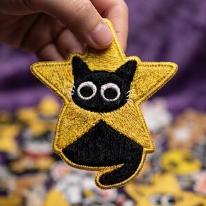 May include: An embroidered patch shaped like a five-pointed yellow star featuring a black cat with large white circular eyes. The cat is positioned in the center of the star with its tail curving along the bottom edge of the yellow star shape.