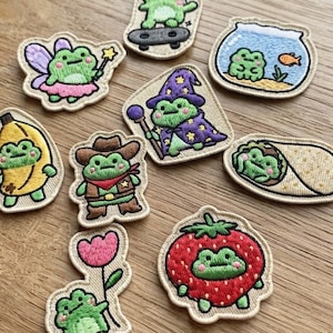 May include: A collection of nine embroidered patches featuring green frogs in various costumes and settings, including a frog on a skateboard, a frog in a banana suit, a frog as a wizard, a frog in a fishbowl, and a frog inside a strawberry.