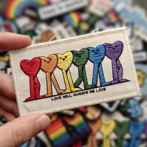 May include: A rectangular embroidered patch on a cream-colored fabric base featuring six heart-shaped figures in red, orange, yellow, green, blue, and purple, each with two legs. The text "LOVE WILL ALWAYS BE LOVE" is printed in black below the hearts.