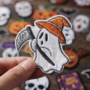 May include: An embroidered patch featuring a white ghost wearing an orange witch hat and holding a silver scythe. The patch has a white border and is held up against a background of various other Halloween-themed embroidered patches.