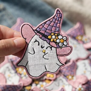 May include: An embroidered patch featuring a white ghost with a smiling face, rosy cheeks, and a small yellow star. The ghost wears a purple and pink checkered witch hat adorned with two white and yellow flowers, all outlined in pink thread.
