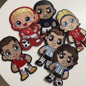 May include: A collection of six embroidered fabric patches featuring cartoon-style soccer players in various team uniforms. The figures include players in red jerseys with numbers 9 and 23, a dark blue jersey with number 10, and light blue striped kits.