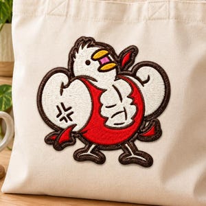 May include: A cream-colored canvas tote bag featuring a large embroidered patch of a muscular white chicken with a red chest, yellow beak, and dark brown outlines. The chicken has a flexed pose with visible muscle definition and a small angry symbol.