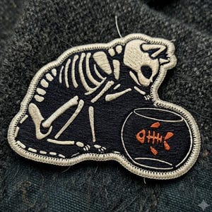May include: An embroidered patch featuring a black cat skeleton sitting beside a round fishbowl. The cat skeleton is stitched in off-white thread against a black background. Inside the fishbowl, an orange fish skeleton is clearly visible.