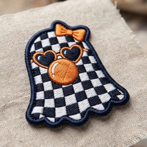 May include: An embroidered patch shaped like a ghost with a black and white checkered pattern. The ghost wears orange heart-shaped sunglasses, has an orange bubble gum bubble in front of its face, and features a small orange bow on its head.