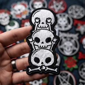 May include: An embroidered patch featuring three stacked white skulls with black details. The top skull covers its eyes, the middle skull covers its ears, and the bottom skull covers its mouth, all outlined in black thread on a dark background.