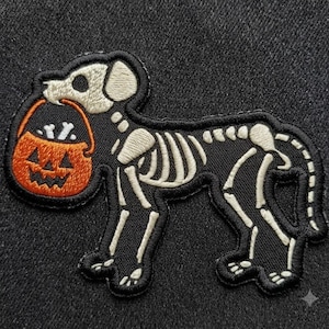 May include: An embroidered patch featuring a dog skeleton in white thread on a black background. The dog is depicted in profile, holding an orange jack-o'-lantern bucket in its mouth, which contains small white bone shapes inside the pumpkin.