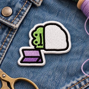 May include: An embroidered patch featuring a green frog character with a simple black line face, partially covered by a large white blanket. Below the frog is a small, rectangular purple and lavender striped book with a black border.
