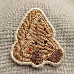 May include: An embroidered patch shaped like a Christmas tree cookie with a light brown base, a cream-colored border, and dark brown feet. The design features a smiling face with black eyes and small red and green embroidered dots as decorations.