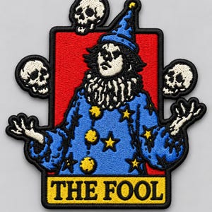 May include: An embroidered patch featuring a figure dressed in a blue wizard-style costume with yellow stars and pom-poms, standing before a red rectangular background. Three white skulls float around the figure, and the text THE FOOL is at the bottom.