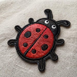 May include: An embroidered ladybug patch featuring a bright red circular body with six black spots. The head and legs are stitched in black thread, and the eyes are white with black pupils. The design is outlined in black thread on a textured fabric.