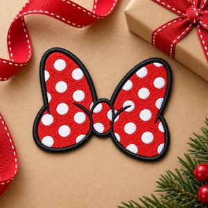 May include: A red bow-shaped embroidered patch featuring a pattern of white polka dots. The patch has a thick black stitched border outlining the shape of the bow, which consists of two large loops and a small central knot.