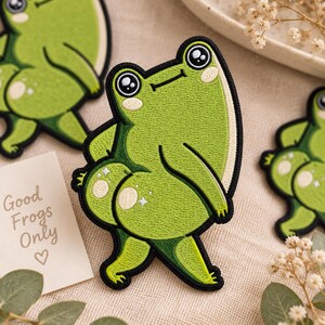 May include: An embroidered patch featuring a cartoon frog with large eyes, light green skin, and dark green accents. The frog is depicted in a standing pose with its back turned, showcasing rounded features with small white star-shaped highlights.