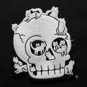 May include: An embroidered patch featuring a white skull design on a black background. The skull is covered in multiple small white cat figures, with cats positioned on the cranium, within the eye sockets, and along the jawline of the skull.