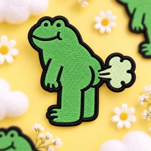 May include: An embroidered patch featuring a bright green frog standing in profile with a black outline. The frog is depicted with a light green cloud shape emitting from its rear, suggesting a flatulent action. The design is clean and graphic.
