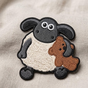 May include: An embroidered patch featuring a black and white sheep character with large eyes and floppy ears, holding a small brown teddy bear. The sheep has a cream-colored woolly body, black face, and black limbs, with a black border outlining the design.